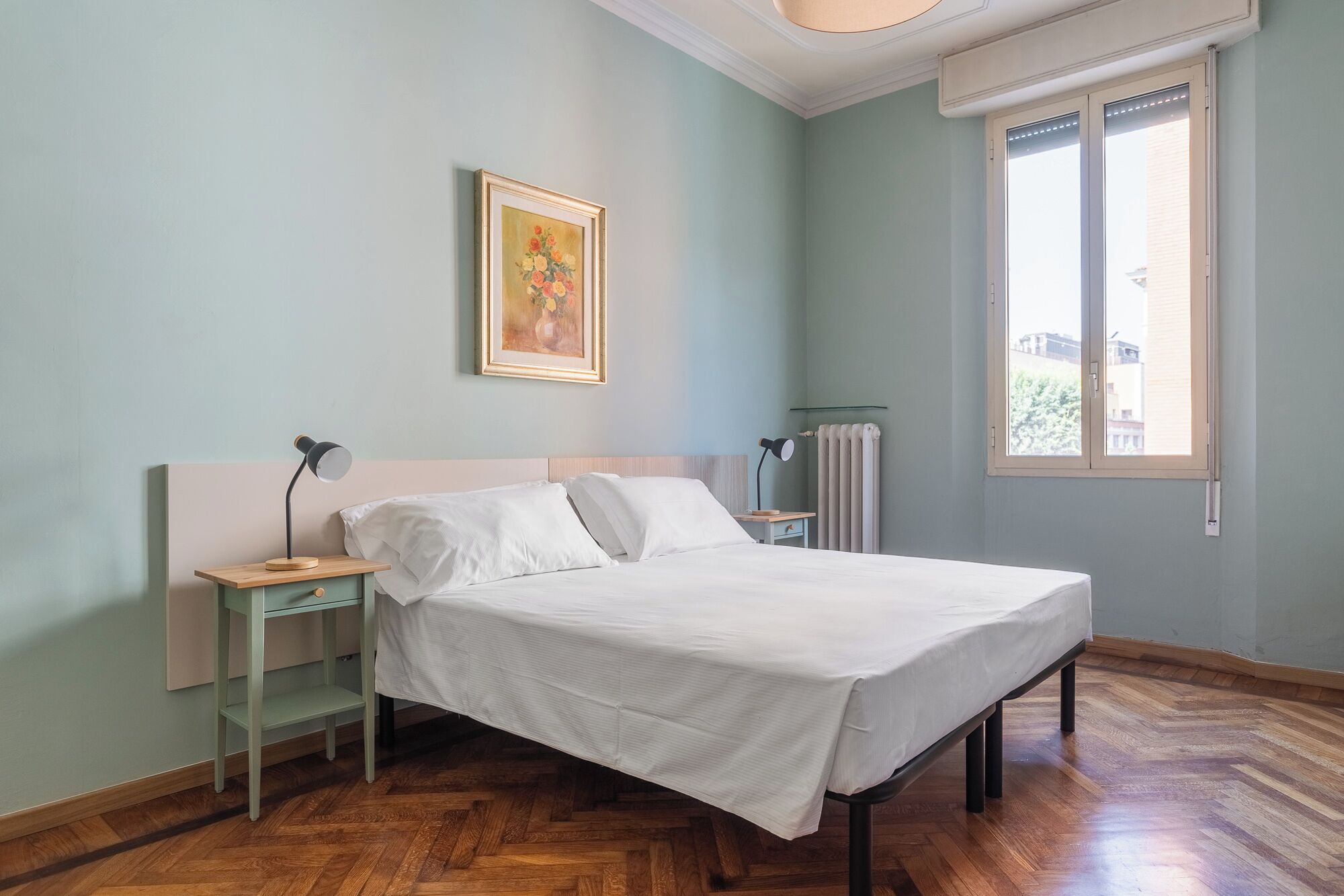 A bedroom with a bed, two bedside tables, a nightstand, a radiator, and a window.