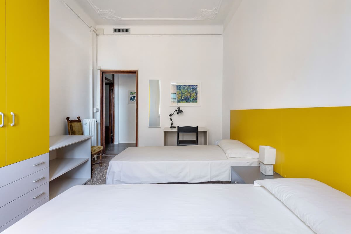 Une chambre d’hôtel avec deux lits, un bureau, une chaise, un radiateur et un tableau au mur.