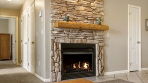 Une cheminée en pierre avec un manteau en bois, un feu qui crépite à l’intérieur, et un couloir menant à une porte.