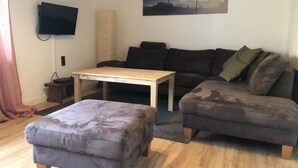A living room with a sofa, ottoman, coffee table, and a flat-screen TV mounted on the wall. - Vacation home Beulskopf - relaxing in nature (Heupelzen)