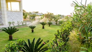 A patio with a white plastic chair and a glass table, surrounded by various plants and a building in the background. - Lydia Apartment (Stavros)
