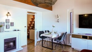 Een eethoek met een witte tafel gedekt voor vier personen, omringd door stoelen. Links is een open haard te zien, en rechts hangt een televisie aan de muur.
