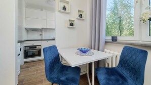 A modern kitchen with white cabinets, a white table with blue chairs, and a window with a view of trees. - Compact Studio | Close to Park, Pet Friendly (Wroclaw)