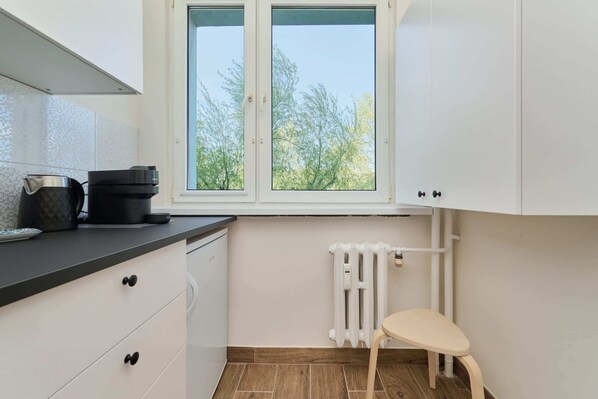 Une cuisine avec des meubles blancs, une fenêtre donnant sur des arbres, un radiateur et un petit tabouret.