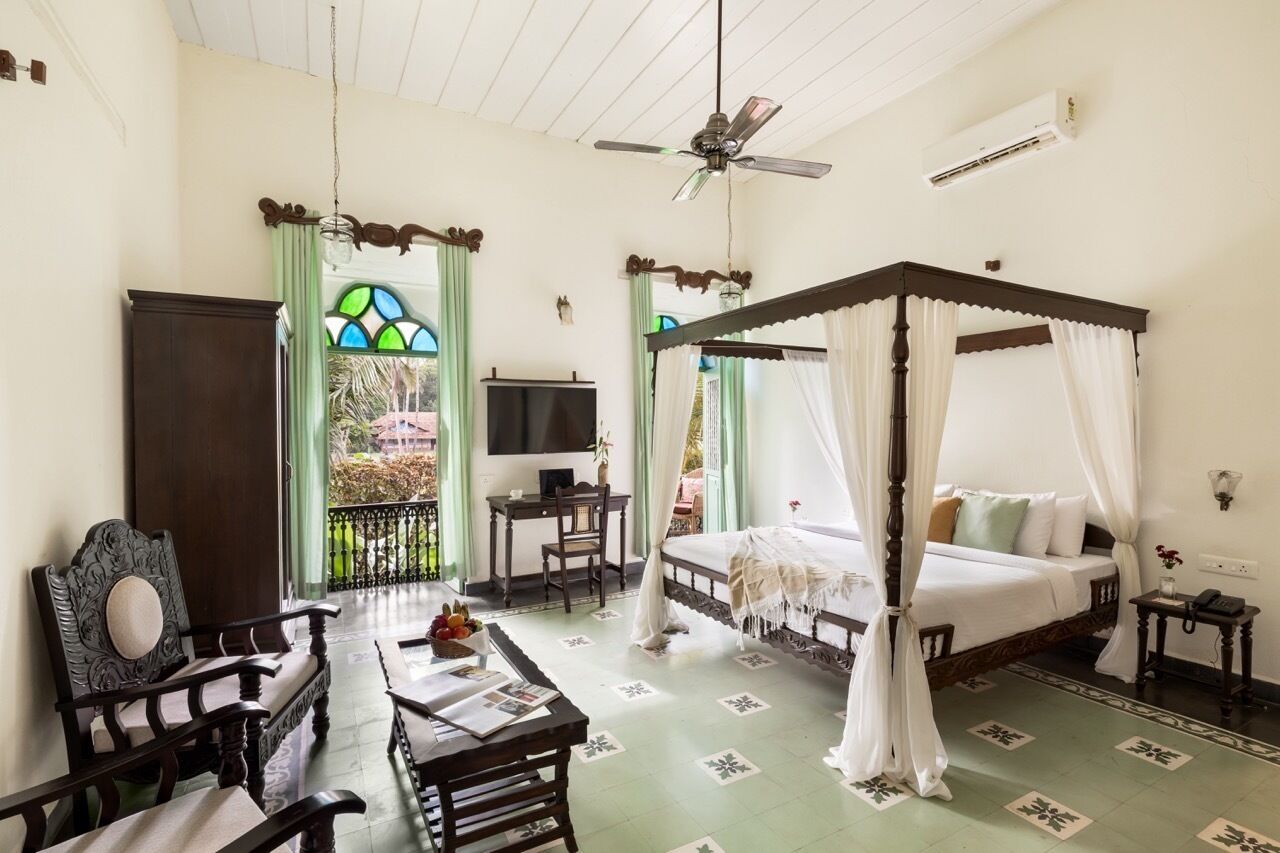 A room with a four-poster bed, a desk with a chair, a television, and a balcony with a view of greenery.