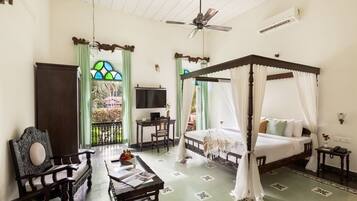 A room with a four-poster bed, a desk with a chair, a television, and a balcony with a view of greenery.