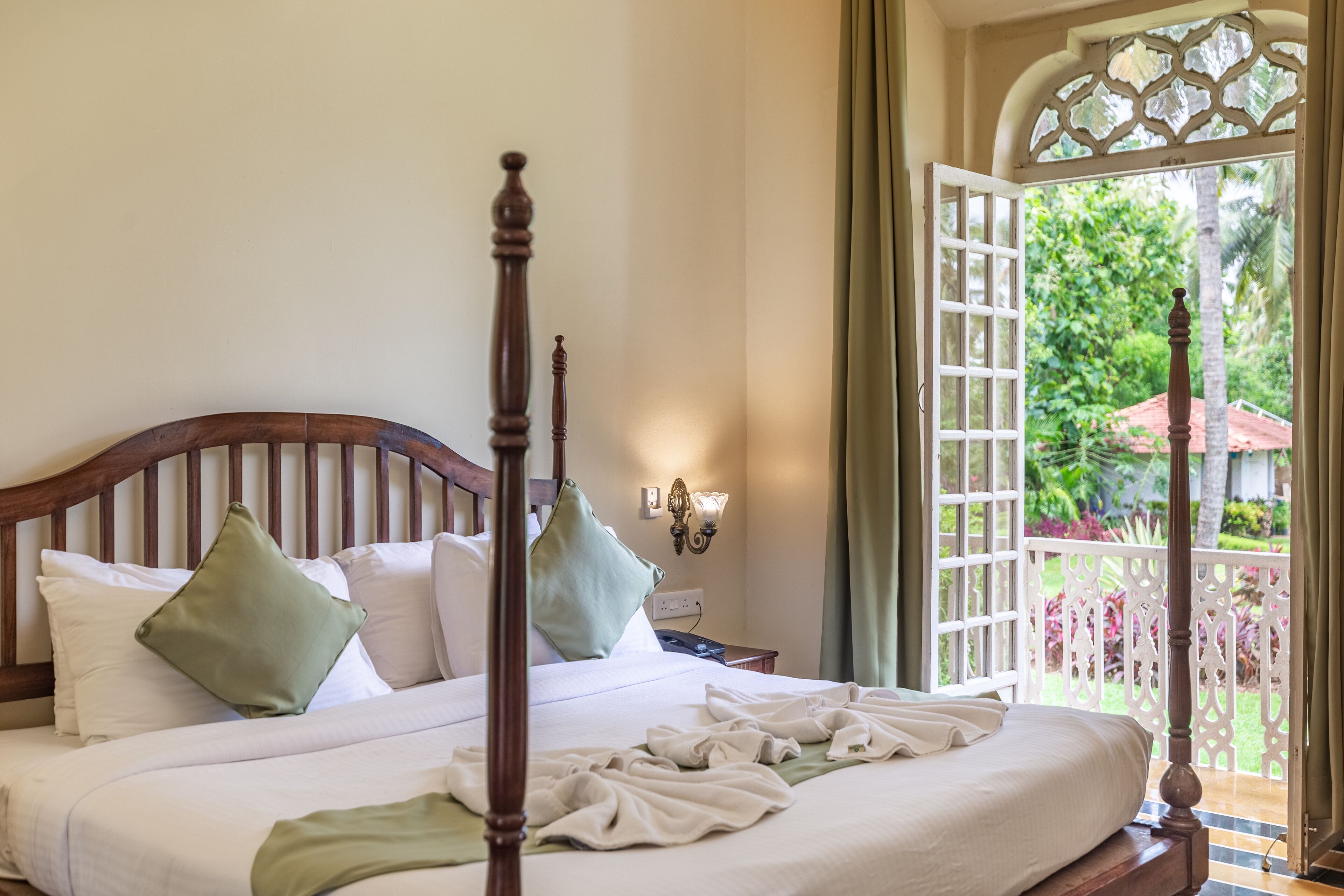 A neatly made bed with a wooden headboard, a nightstand with a lamp, and a view of a lush garden through an open door.