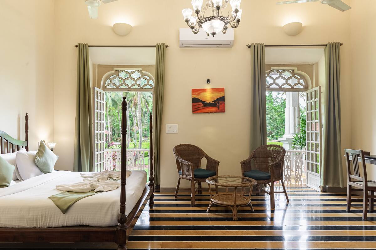 A room with a bed, two wicker chairs, a small round table, and a painting of a sunset.