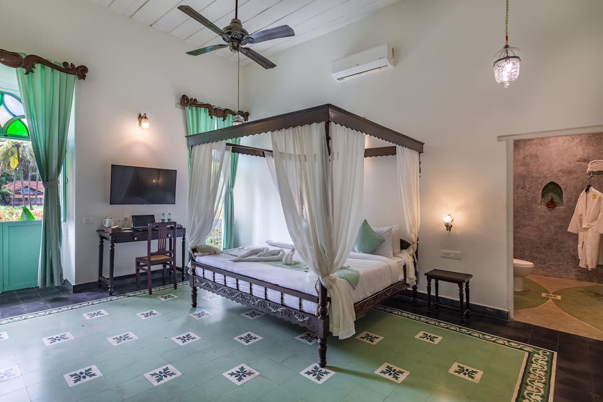 A four-poster bed with white bedding, a desk with a laptop, a television, and a green patterned floor.