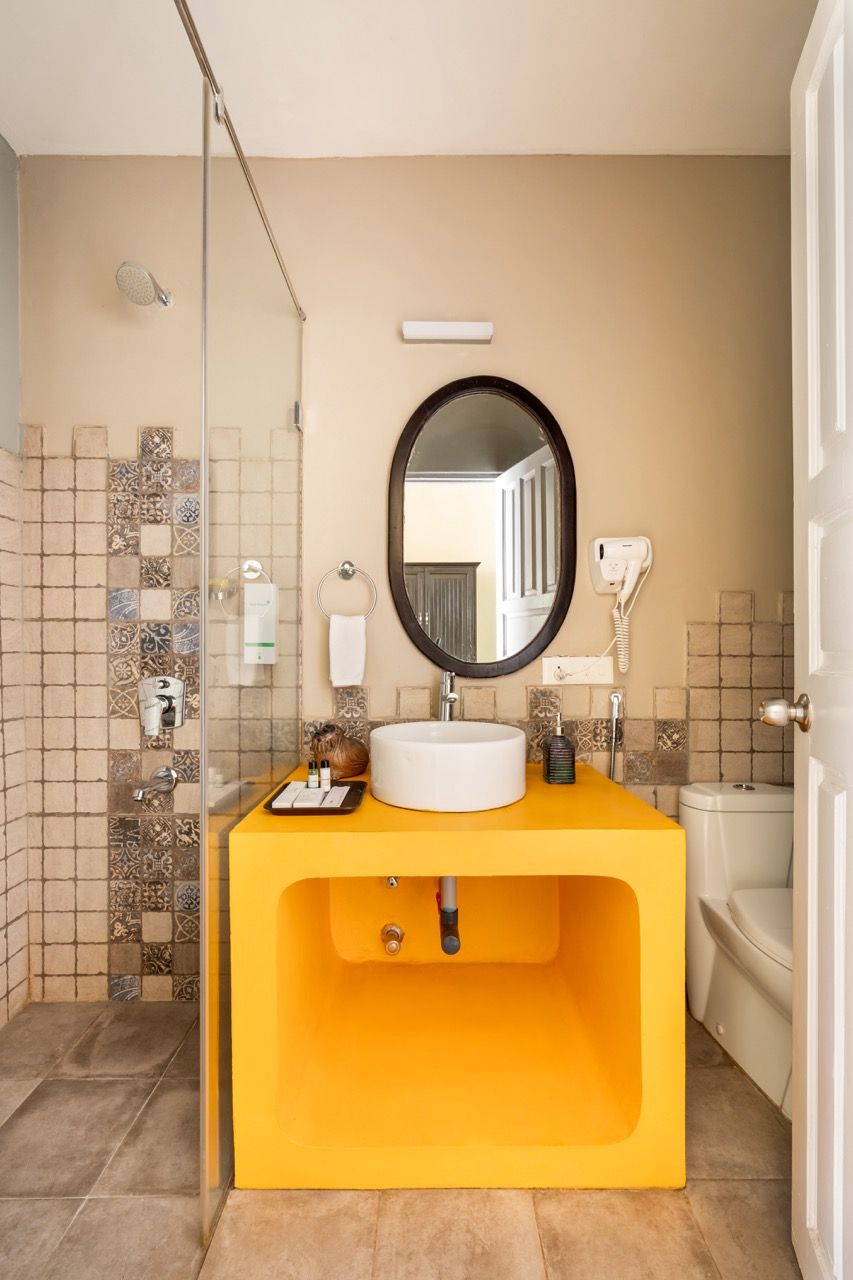 Un baño con un mueble de tocador amarillo, un espejo redondo, un lavamanos blanco, un cerramiento de ducha de vidrio y un inodoro.