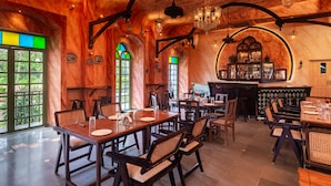 A restaurant with wooden tables and chairs, a bar area, and large windows with green glass panes. - Silva Heritage Resort Goa (Colva)