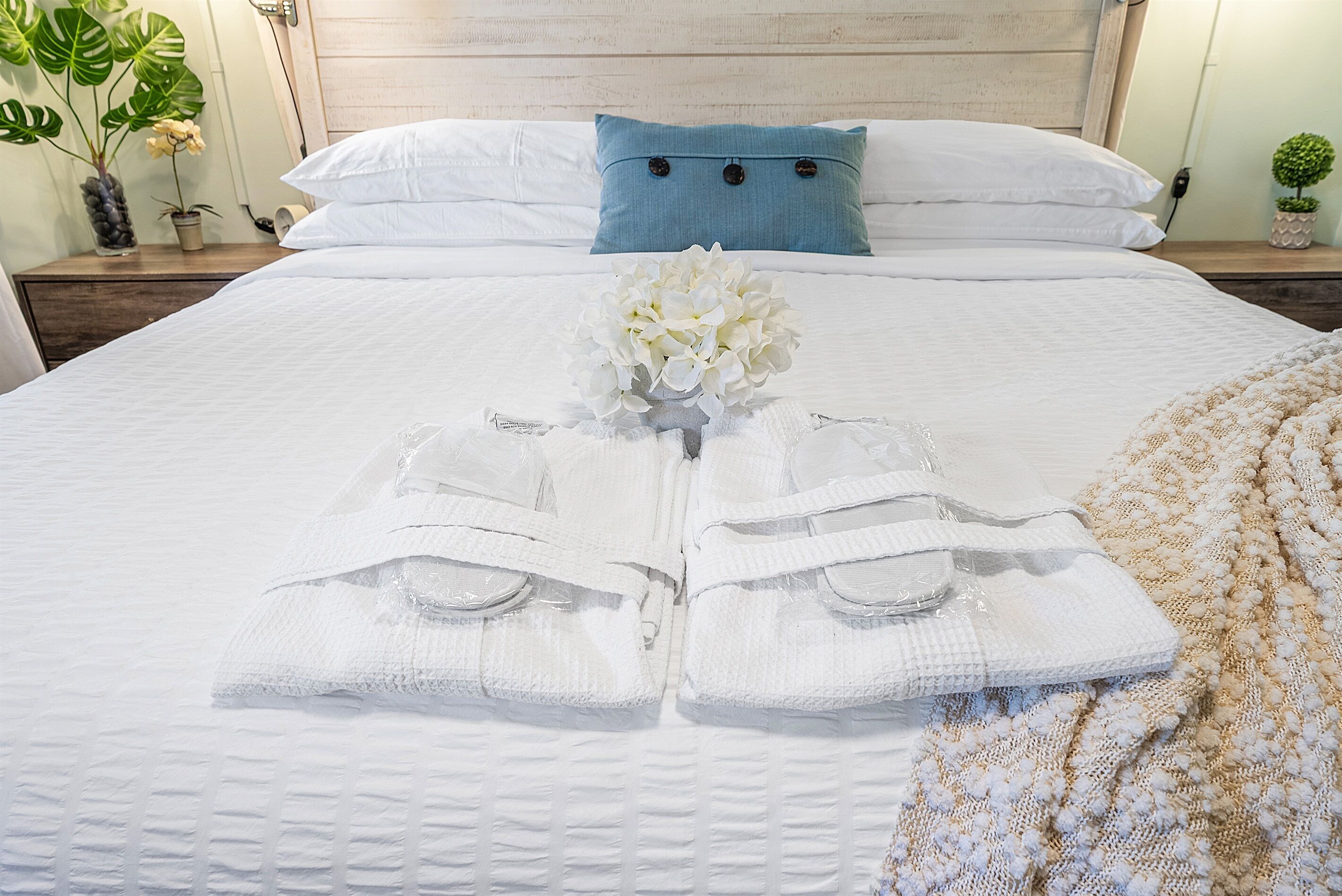 Una cama bien hecha con sábanas blancas, un almohadón azul y un arreglo floral blanco.