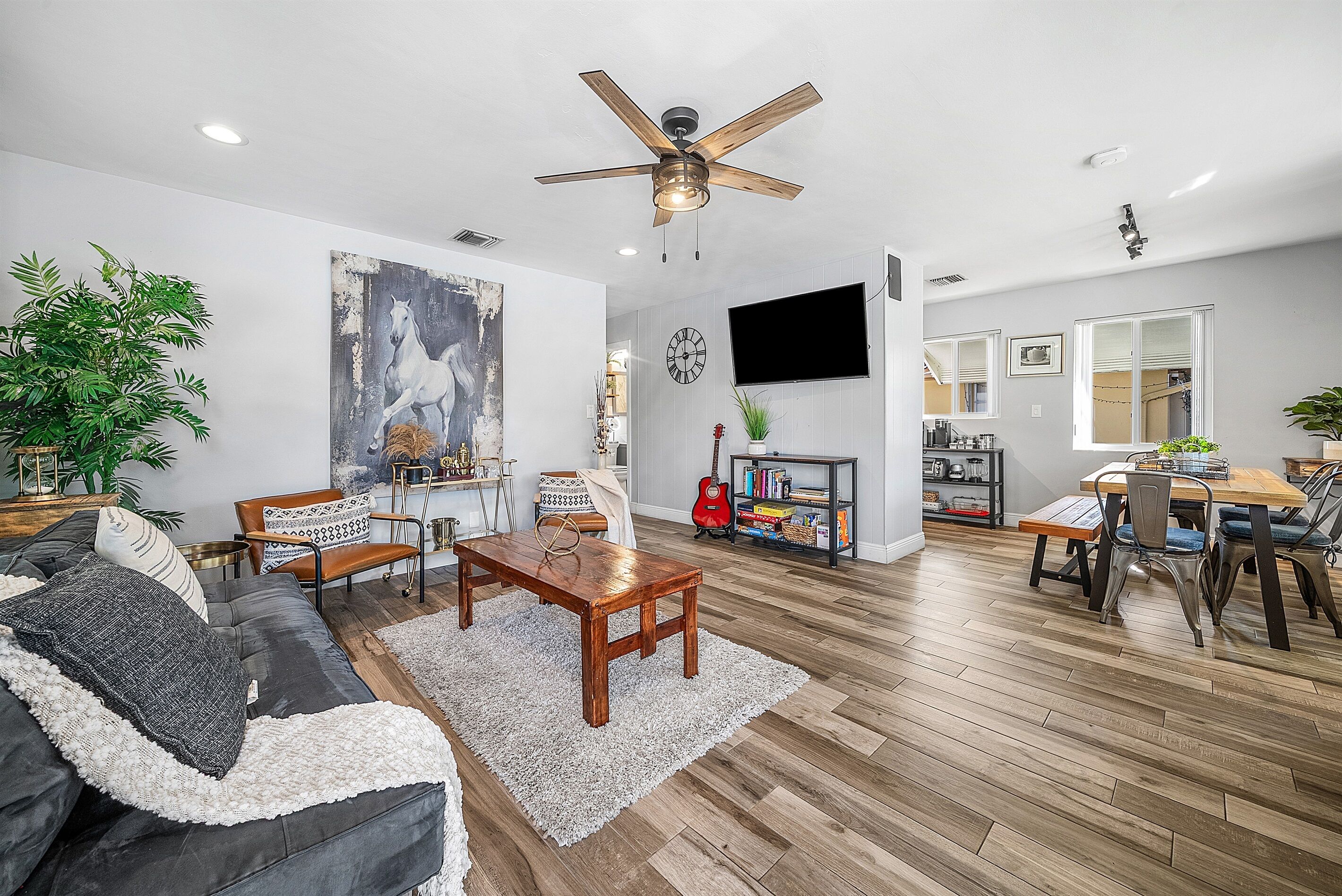 A spacious living room with a wooden floor, a ceiling fan, a large painting, a dining area with a table and chairs, and a TV mounted on the wall.