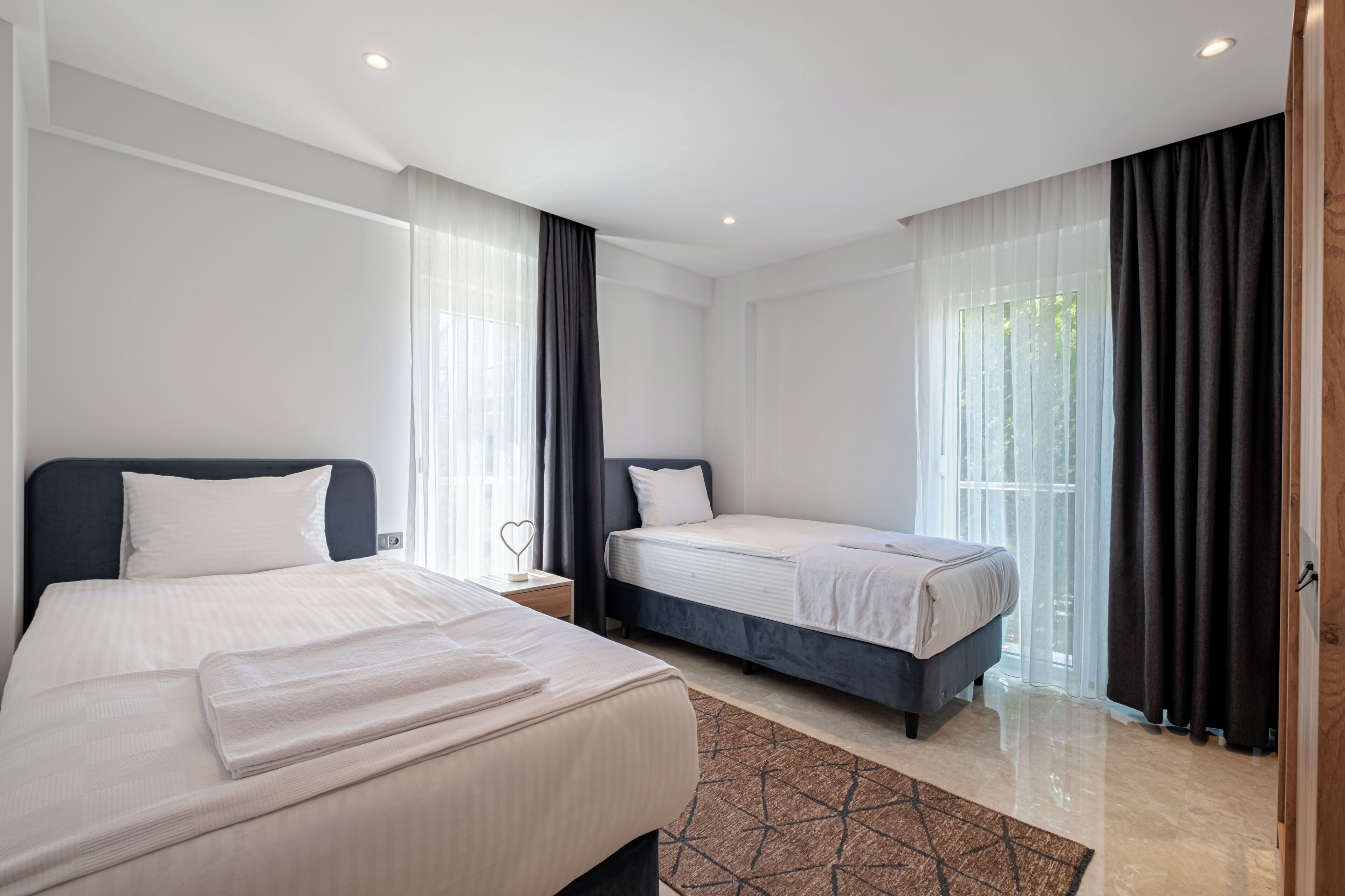 A modern hotel room with two beds, a large window with sheer curtains, and a clean, minimalist design.