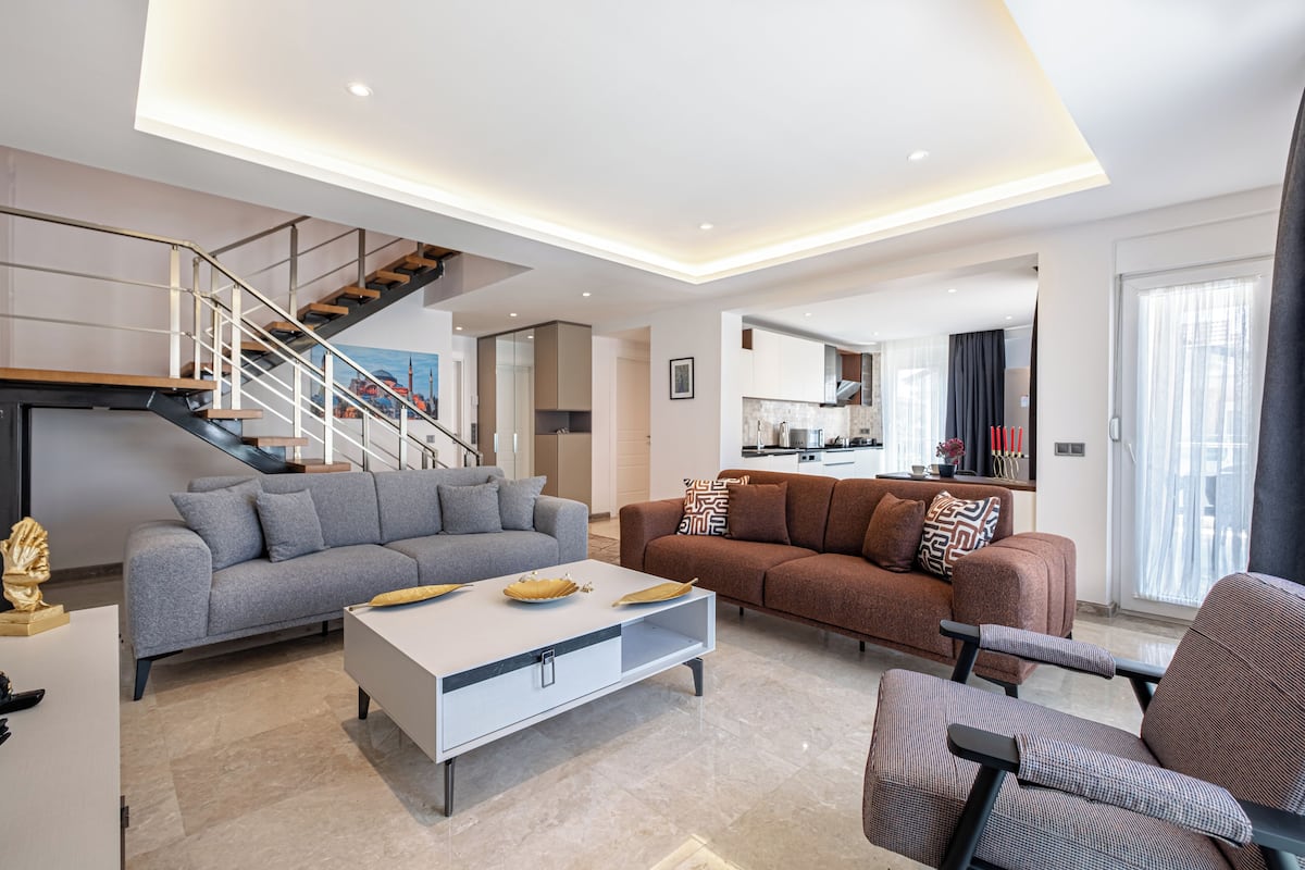 A modern living room with a grey sofa, a brown sofa, a white coffee table, and a patterned armchair. There is a staircase with a railing leading to an upper level.