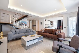 Una sala moderna con un sofá gris, un sofá marrón, una mesa de centro blanca y un sillón estampado. Se observa una escalera con barandilla que lleva a un nivel superior.