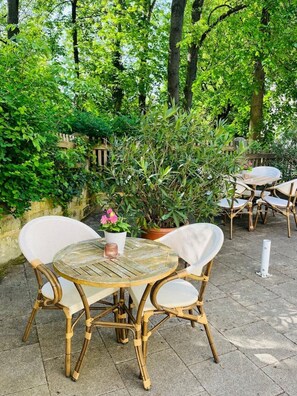 An outdoor seating area with wicker chairs and a round table, surrounded by lush greenery and a wooden fence.