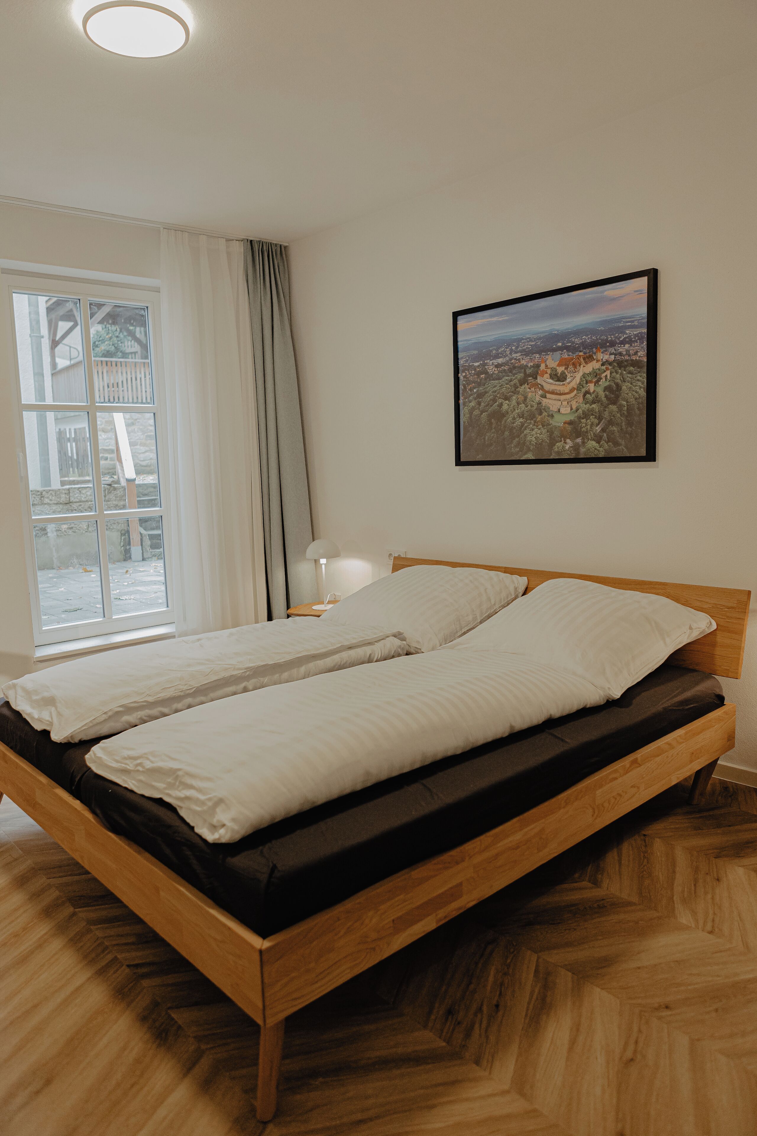 Ein Schlafzimmer mit einem hölzernen Bett, einem Fenster mit Vorhängen, einer Nachttischlampe und einem gerahmten Bild an der Wand.