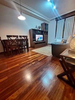 A modern living room with a wooden floor, a TV on a stand, a sofa, a dining table with chairs, and a ceiling light.