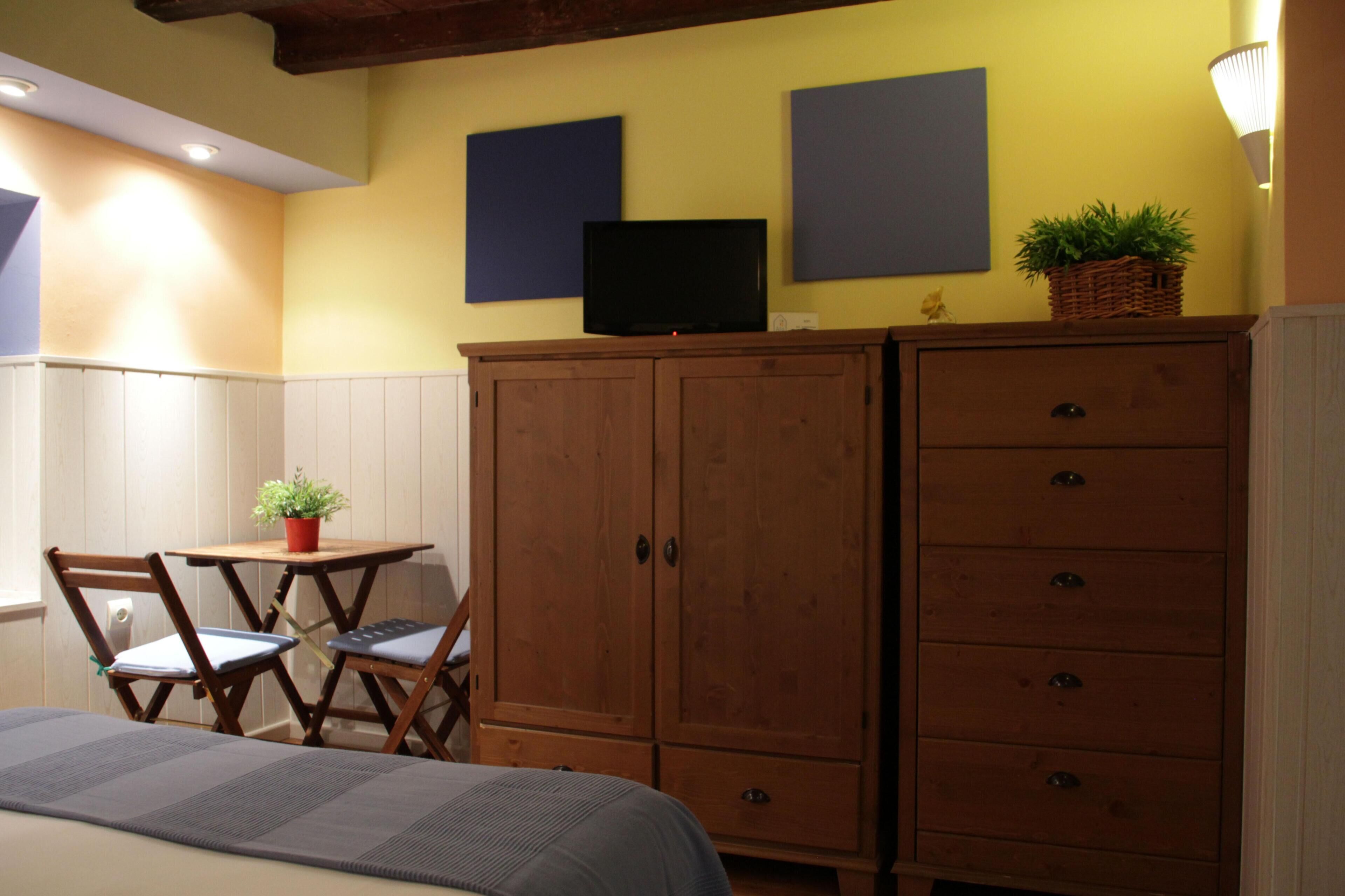 A room with a bed, wooden wardrobe, a small table with a plant, two chairs, and a television on the wall.