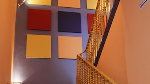 Un escalier en bois, agrémenté d’une balustrade dorée, mène à un palier dont les murs sont ornés de panneaux carrés aux couleurs vives.