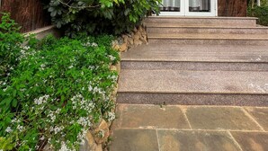 A house with a stone staircase leading to a glass door, surrounded by lush greenery and pink flowers. - 3 Tang May Home & Coffee (Xuan Truong - Da Lat)
