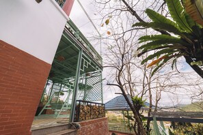 A building with a red brick wall, a green metal structure, and a tree with broad leaves. - 3 Tang May Home & Coffee (Xuan Truong - Da Lat)