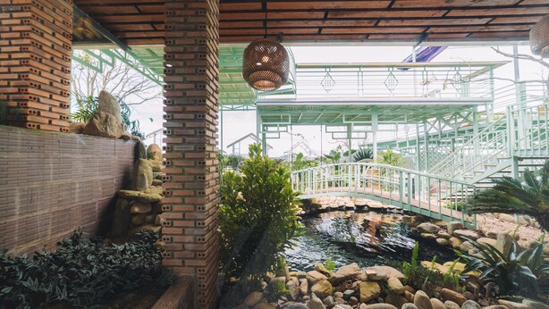 A brick wall with a small garden area, a wooden bench, and a metal bridge over a water feature. - 3 Tang May Home & Coffee (Xuan Truong - Da Lat)