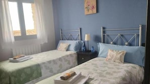 A room with two single beds, each with a green bedspread and white pillows. There is a small table with a lamp and two water bottles. A window with white curtains is visible, and a painting hangs on the wall.