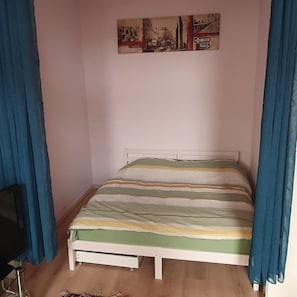 A small, single-bed room with a striped bedspread, a white headboard, and a wall-mounted artwork depicting a cityscape.