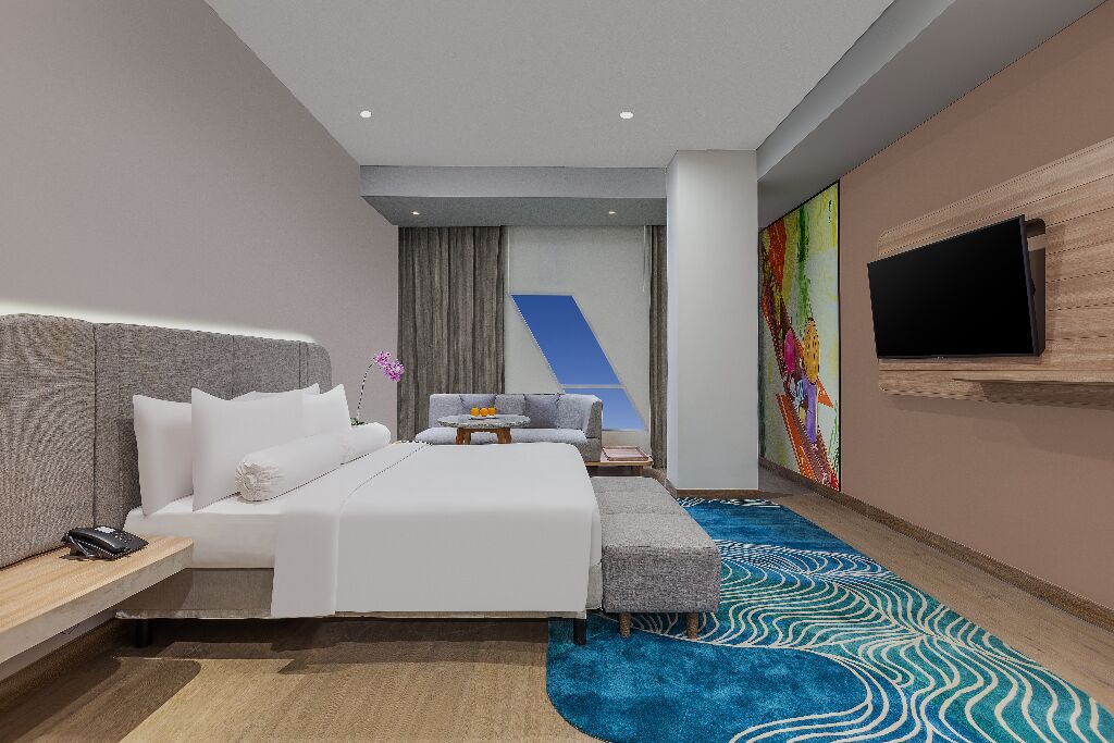 A modern hotel room with a large bed, a sofa, a TV mounted on the wall, and a colorful abstract painting.