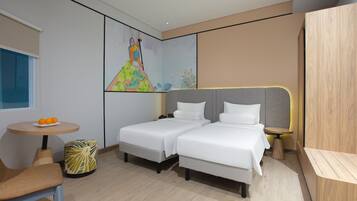 Quarto de hotel moderno com duas camas, uma mesinha redonda com laranjas, uma cadeira e um quadro colorido na parede.