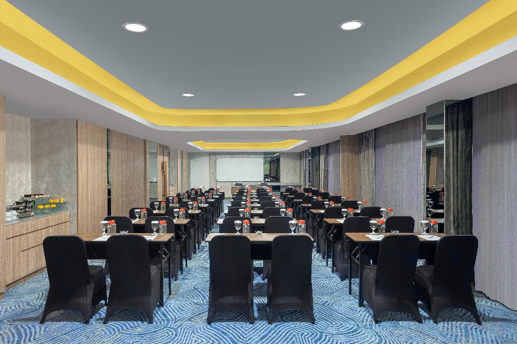 Un salón de conferencias con filas de mesas y sillas, una pantalla grande al frente y un diseño moderno de techo.