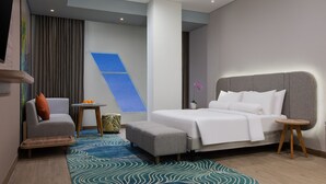 Una habitación de hotel moderna con una cama grande, un sillón gris, una mesita redonda con naranjas, un tapete estampado y un ventanal con un diseño geométrico azul.