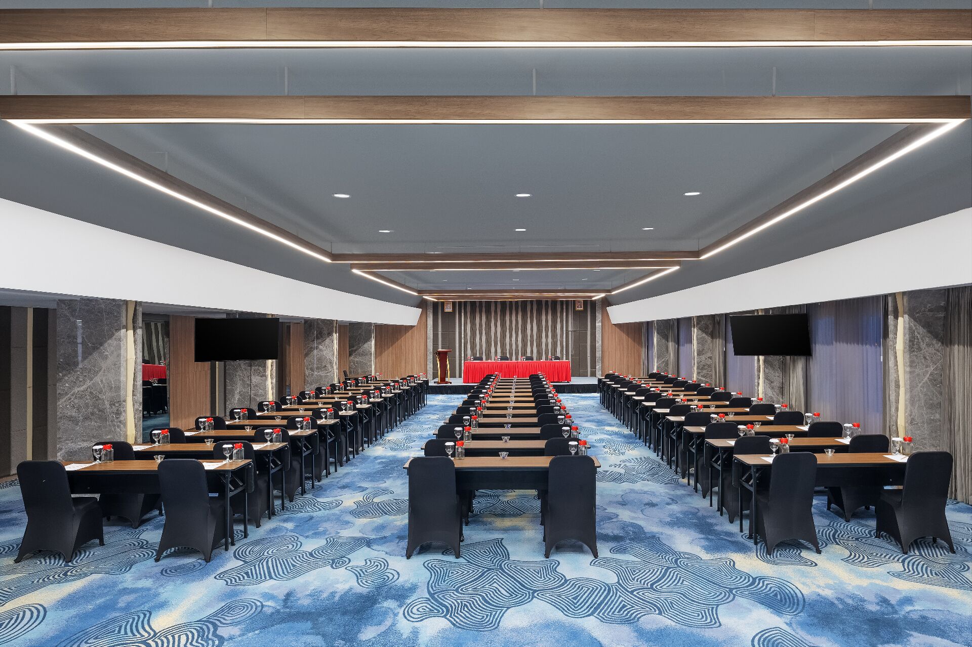 A conference room with rows of tables and chairs, a stage with a red backdrop, and a large screen.