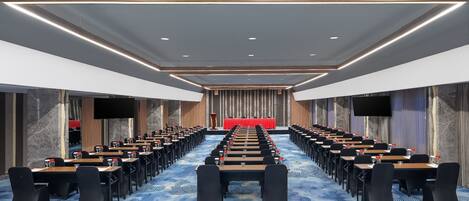 A conference room with rows of tables and chairs, a stage with a red backdrop, and a large screen.
