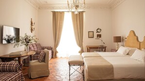 A hotel room with a large bed, a smaller bed, a desk, a chair, a sofa, a TV, and a chandelier. - Casa Pizarro Hotel (Cáceres)