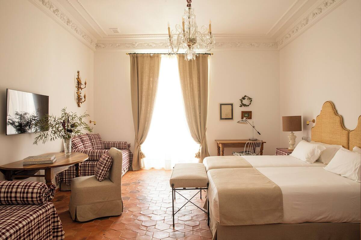 Quarto de hotel com uma cama grande, uma cama menor, uma escrivaninha, uma cadeira, um sofá, uma TV e um lustre.