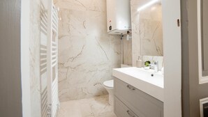 Un baño con pared de mármol blanco, inodoro blanco, lavamanos blanco con espejo y toallero blanco.