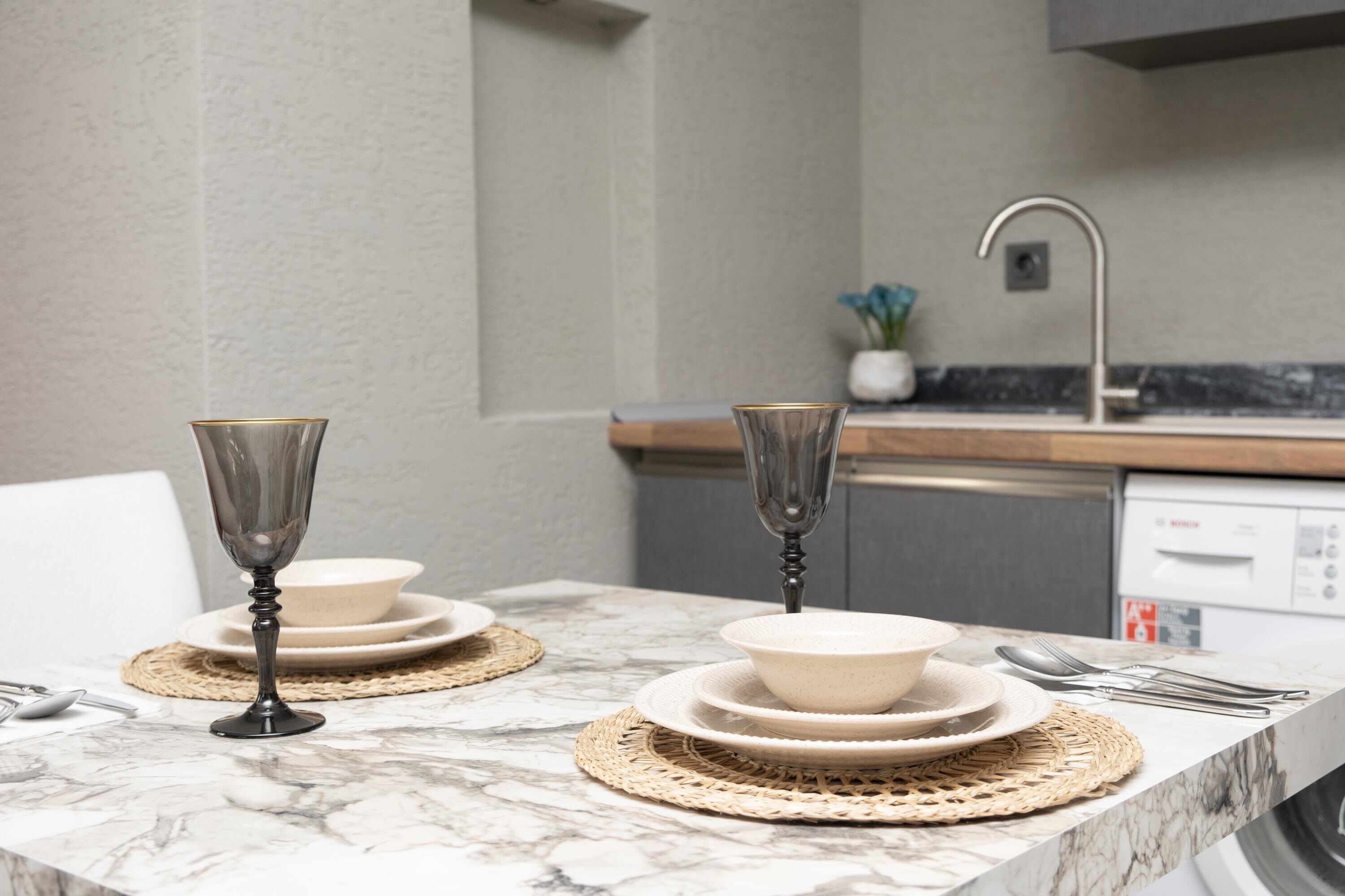 A kitchen with a marble countertop, two wine glasses, stacked plates, and a dishwasher.