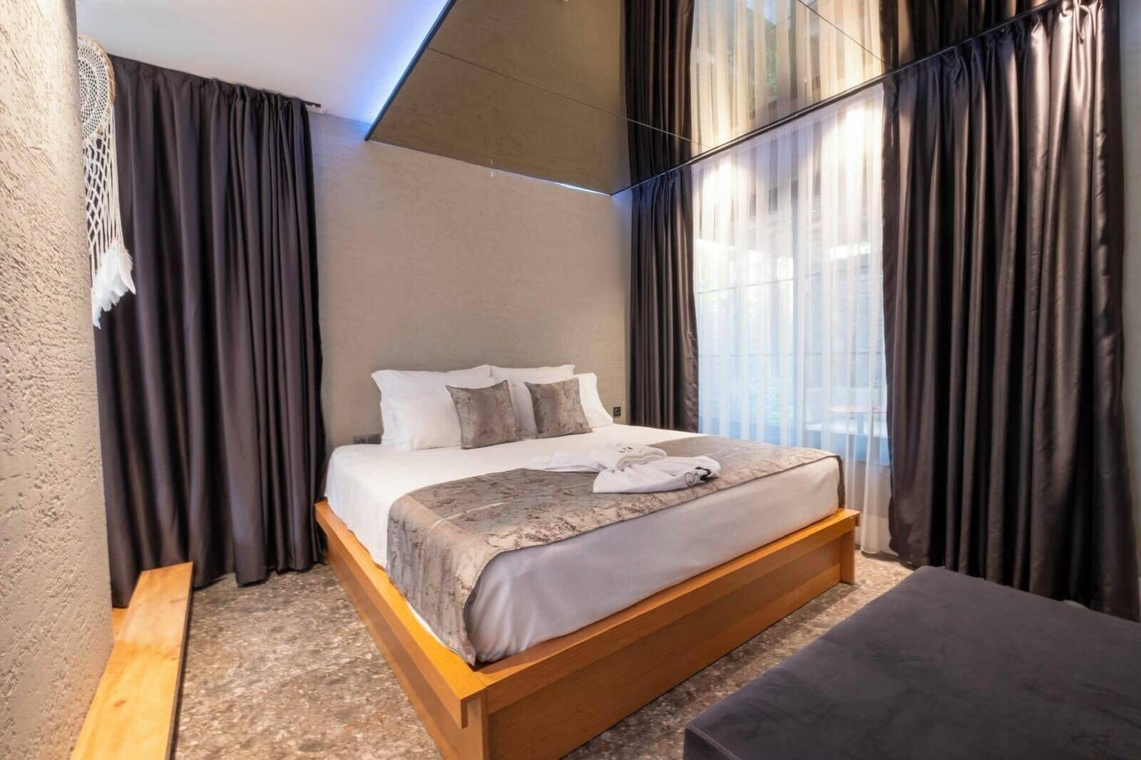 A modern hotel room with a wooden bed, dark curtains, and a view of the outdoors.