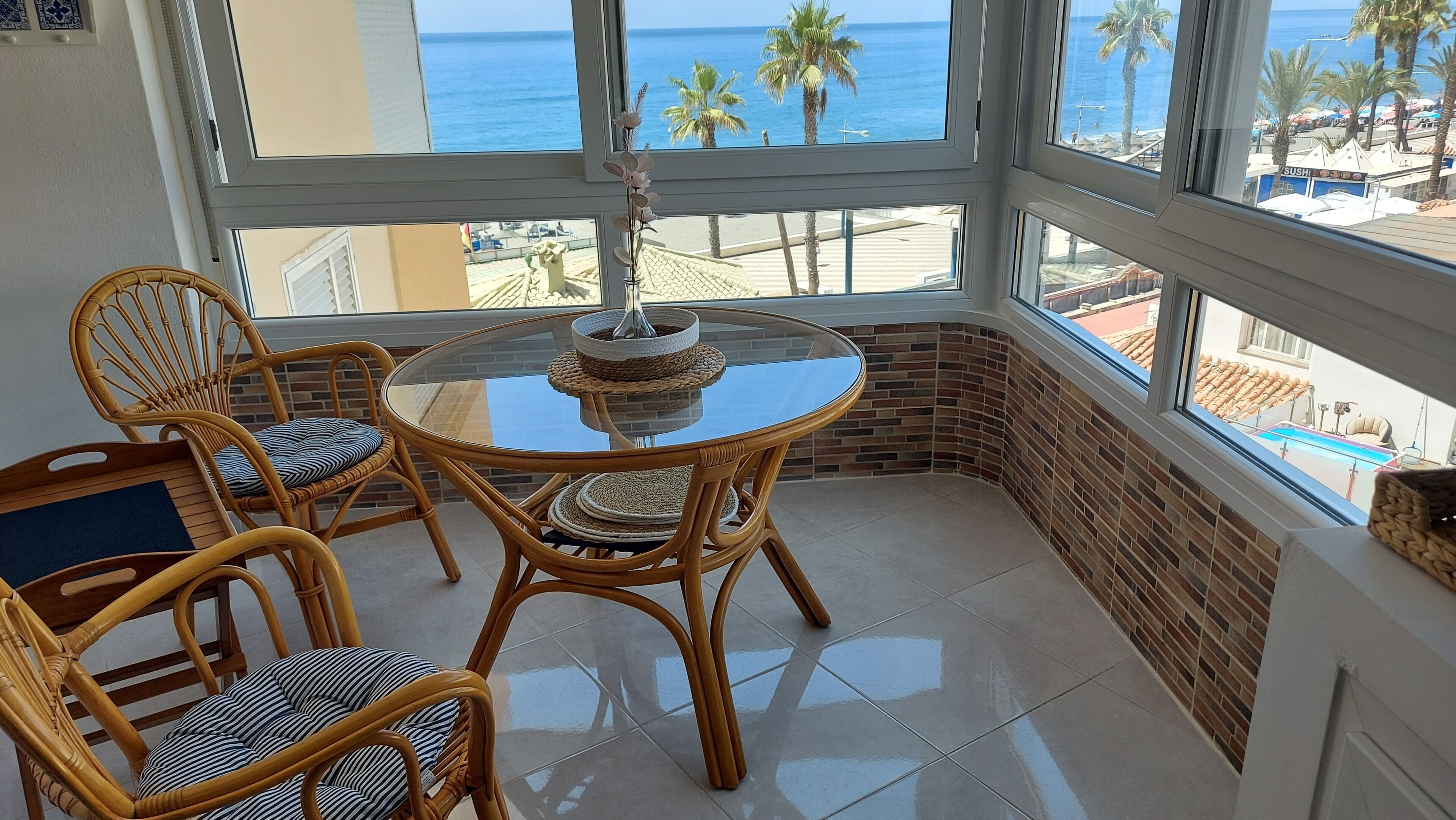 A corner balcony with wicker chairs, a glass table, and a view of the sea and beach.