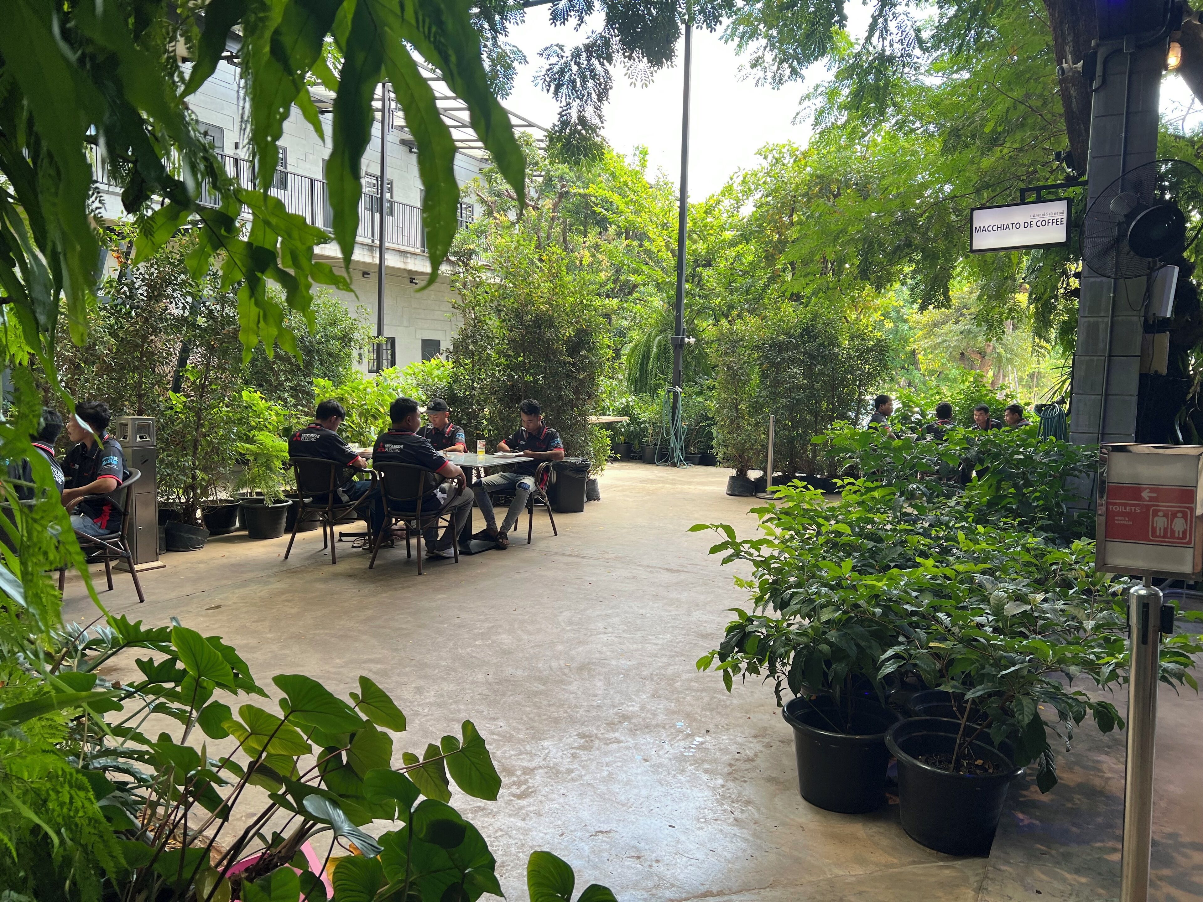 A courtyard with a group of people seated at a table, surrounded by potted plants and a sign indicating a coffee area.
