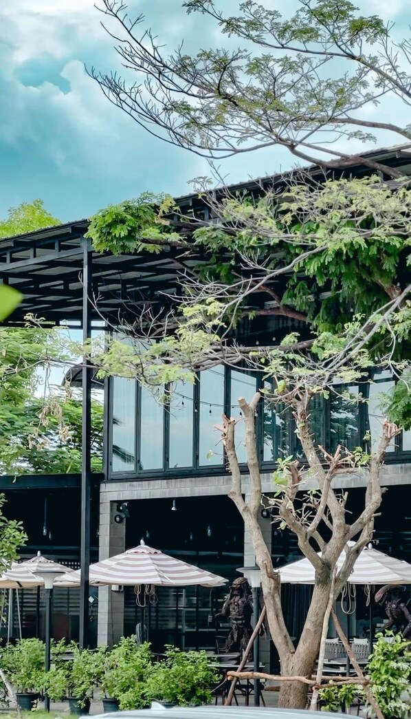 A modern building with a glass facade, outdoor seating area with umbrellas, and a tree in the foreground.