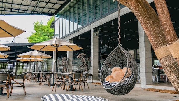 An outdoor seating area with wicker chairs and striped cushions, a hanging chair with a teddy bear, and umbrellas providing shade. - Macchiato Resort Savannakhet (Savannakhet)