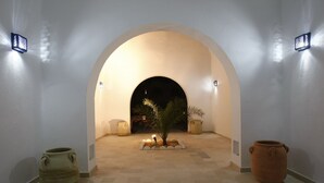 Un pasillo arqueado con una pequeña palmera en el centro, flanqueado por dos grandes jarrones e iluminado por apliques de pared.