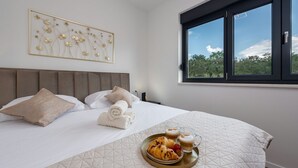 A neatly made bed with a quilted cover, a tray with croissants and coffee, and a window with a view of trees.