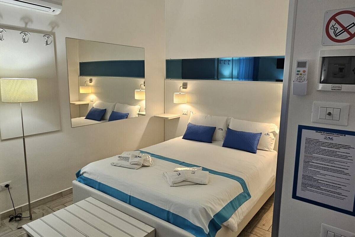 Quarto de hotel com cama, luminárias de cabeceira, espelho, placa de proibido fumar, painel de controle e um quadro de avisos.