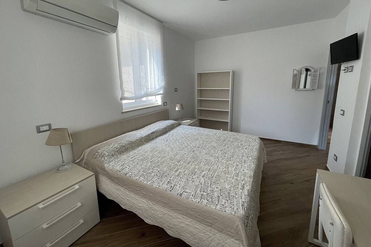 A bedroom with a bed, a nightstand, a lamp, a window with curtains, a built-in shelf, and a wardrobe.
