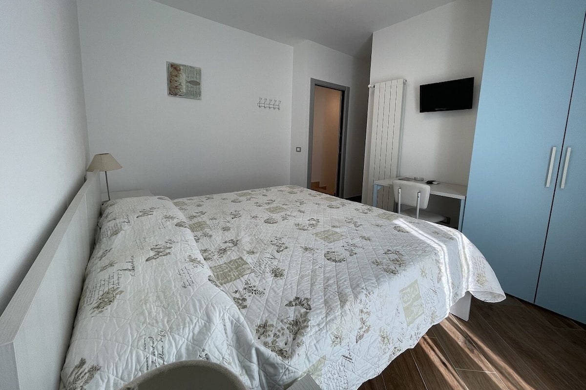 A bedroom with a bed, a desk, a chair, a wardrobe, and a television on the wall.